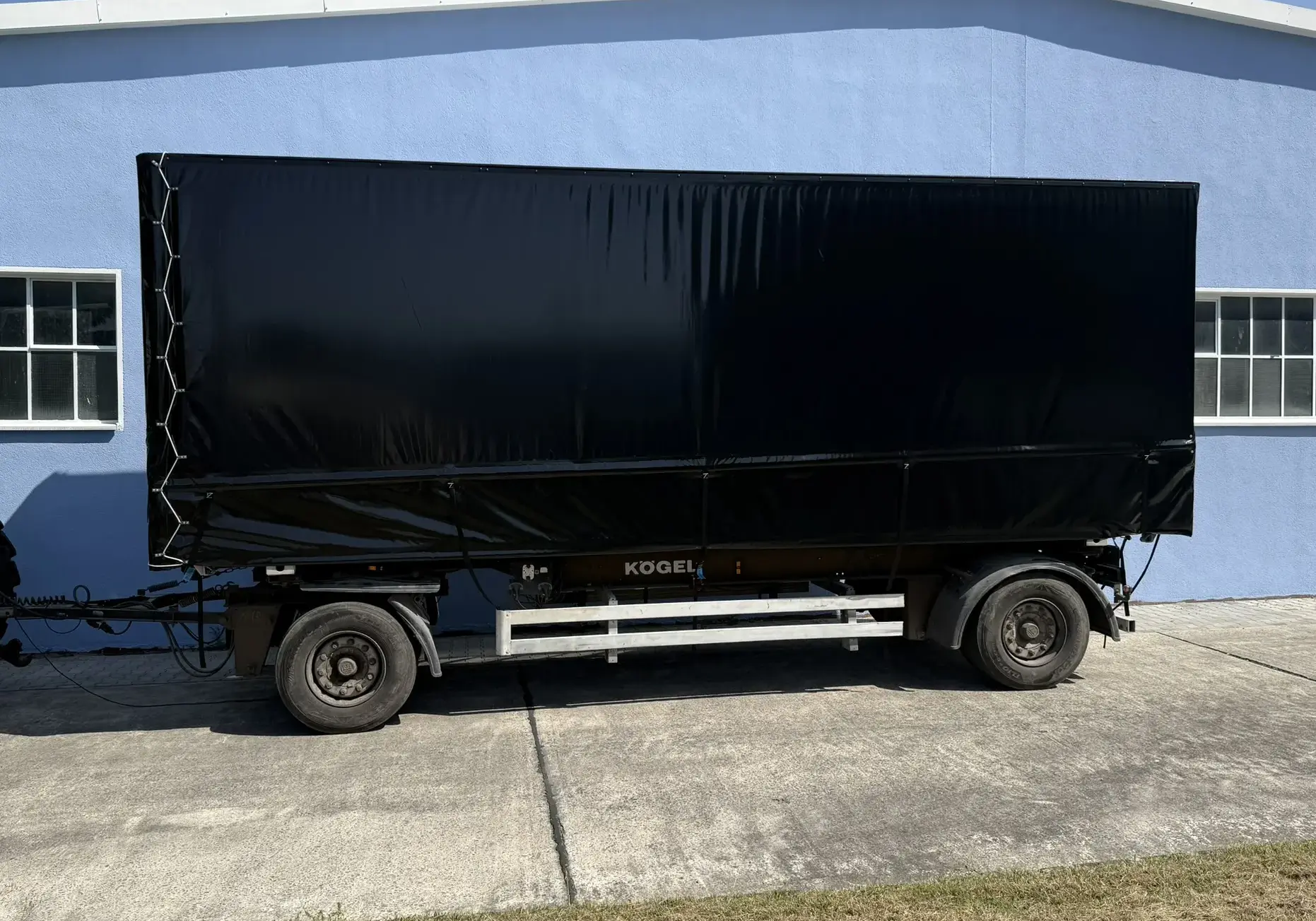 Schwarze Anhängerplane auf einem Kögel-Anhänger, der an einem Gebäude abgestellt ist, mit klaren Linien und einer glatten Oberfläche.